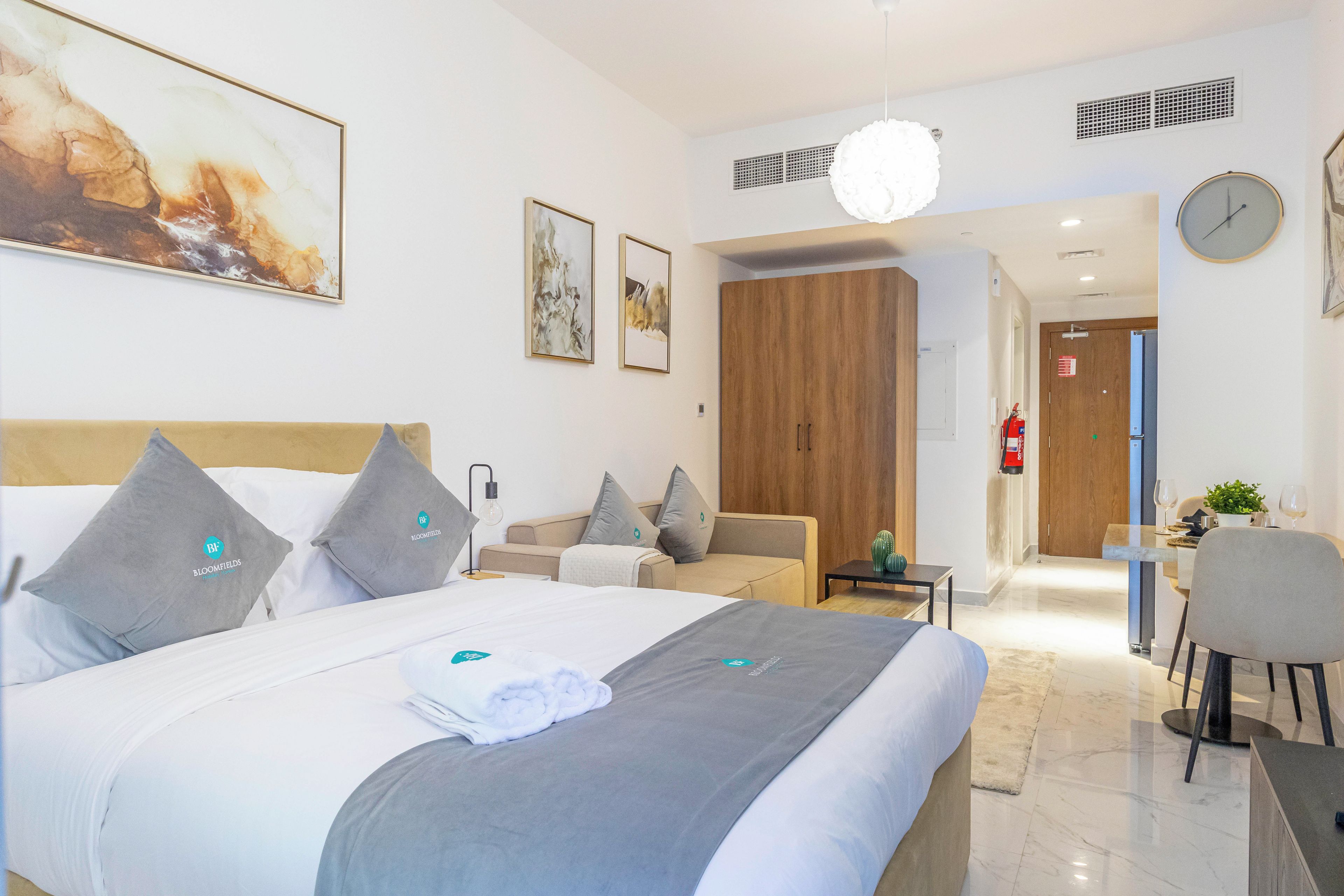 A modern hotel room with a large bed, a sitting area with a sofa, a dining table with chairs, and a kitchen area.