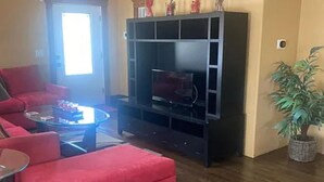 A living room with a red sofa, a glass coffee table, a black entertainment unit, and a potted plant. - Walk to Beach & Festivities All Summer at the Jersey Shore! (Long Branch)