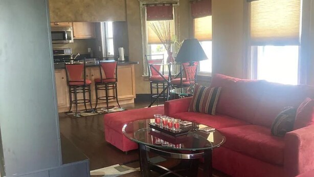 A living room with a red sofa, a glass coffee table, and a kitchen area in the background. - Walk to Beach & Festivities All Summer at the Jersey Shore! (Long Branch)