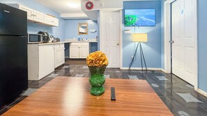 Une cuisine compacte avec des meubles blancs, un réfrigérateur noir, un four à micro-ondes et une cuisinière. On y trouve un vase vert avec des fleurs sur une table en bois, une télécommande et un téléviseur accroché au mur qui diffuse un paysage.