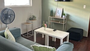A living room with a sofa, a table, a TV mounted on the wall, a fan, and a window with blinds. - Apartment centrally located near Mambo beach Curacao (Willemstad)