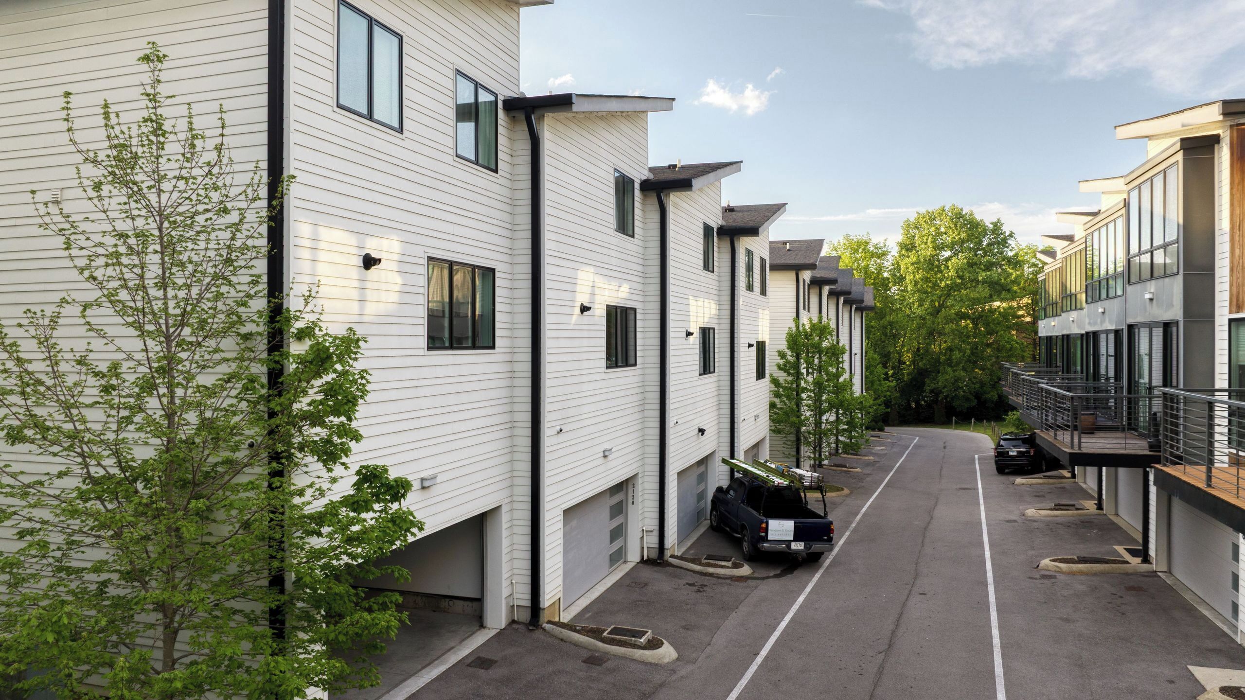 Une rangée d’immeubles modernes et blancs, dotés de nombreuses fenêtres et de balcons, un ciel dégagé et une rue bordée d’arbres.