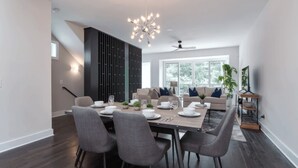 A modern dining area with a large table set for six, surrounded by grey chairs. A contemporary chandelier hangs from the ceiling, and a ceiling fan is visible. The room features dark wooden flooring and a large window with a view of greenery outside. - Luxe 4bdr w/ Balcony & Parking, 10 Mins to Broadway 9th Avenue by Avantstay (Nashville)