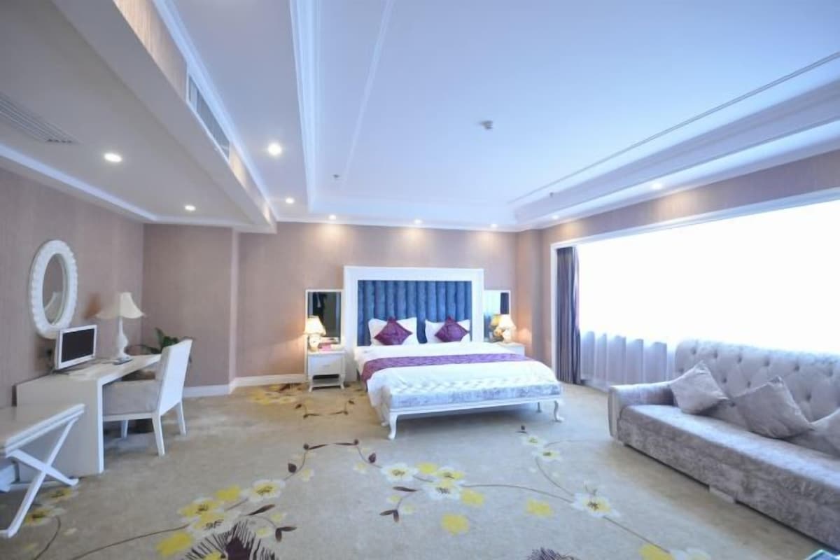 A hotel room with a bed, a sofa, a desk with a chair, and a large window.