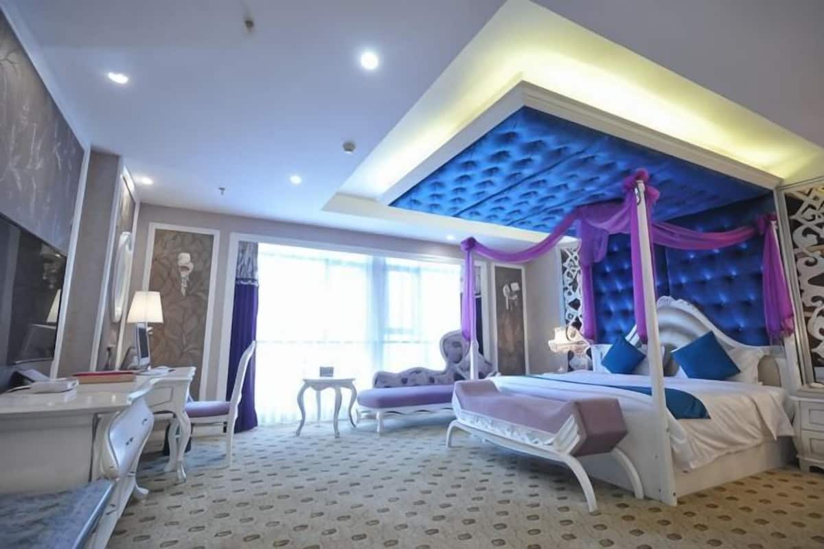 A bedroom with a four-poster bed, a sitting area with a chair, a desk, and a lamp.