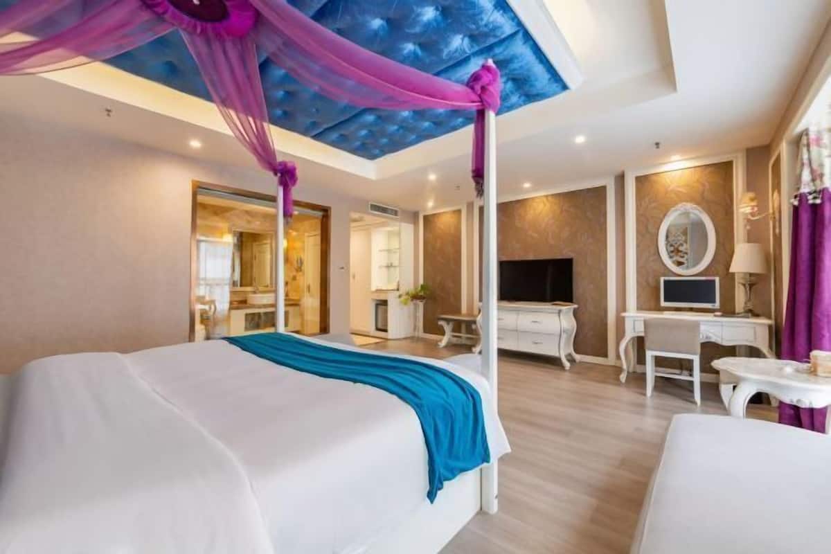 A hotel room with a canopy bed, a desk, a TV, and a bathroom visible through an open door.