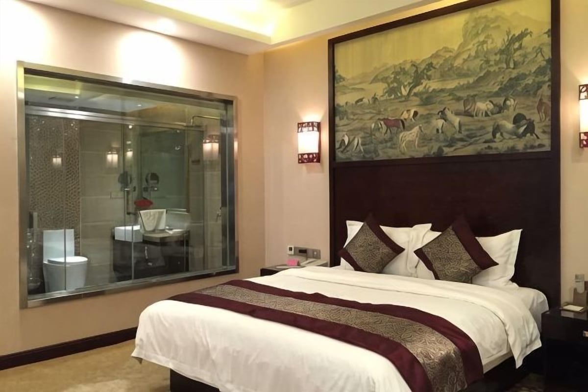 A hotel room with a large bed, a glass shower enclosure, a painting on the wall, and a nightstand with a lamp.