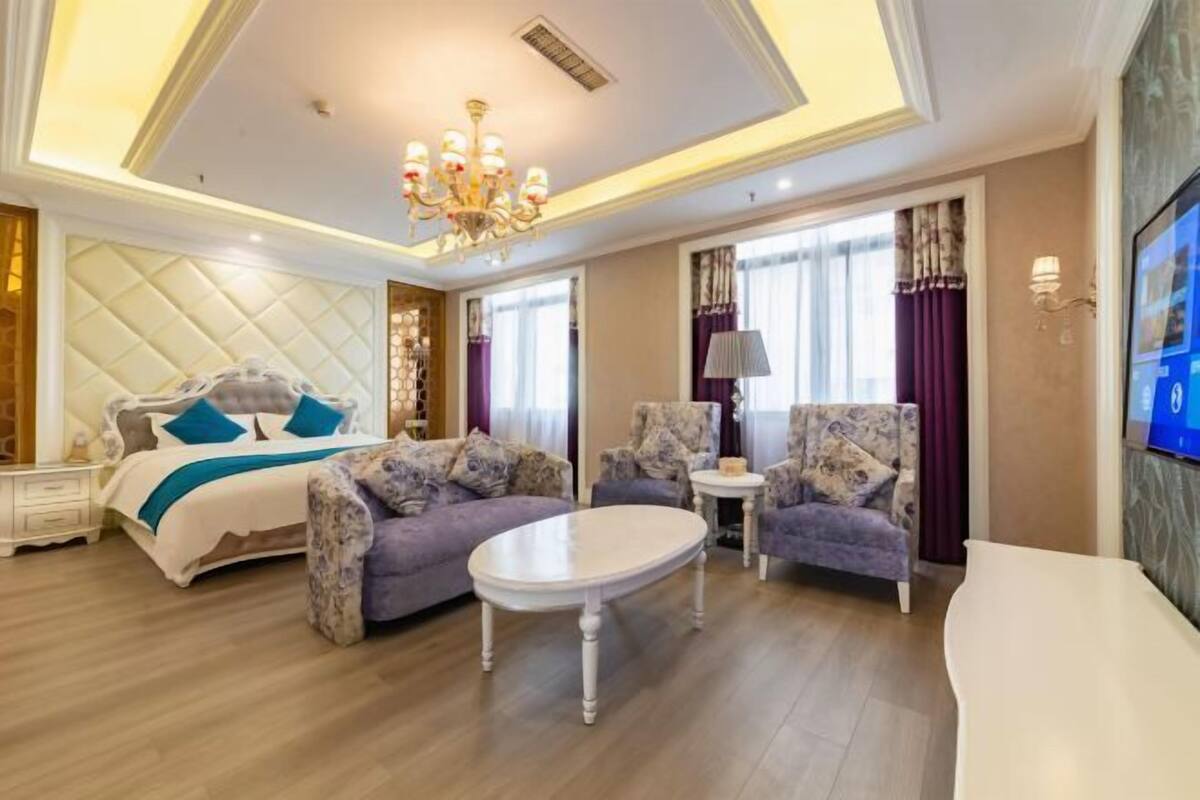 A spacious hotel room with a large bed, two armchairs, a small round table, and a TV mounted on the wall.