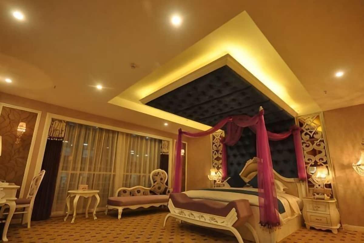 A bedroom with a canopy bed, a sitting area with chairs, a small table, and a large window with curtains.