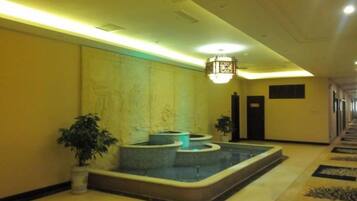 A hotel lobby with a reception desk, a seating area with a fountain, and a hallway with doors.