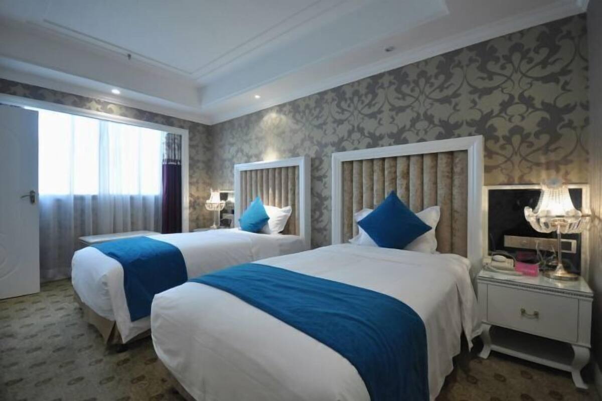 A hotel room with two beds, each with a blue and white bedspread, a white nightstand with a lamp, and a patterned wallpaper on the walls.