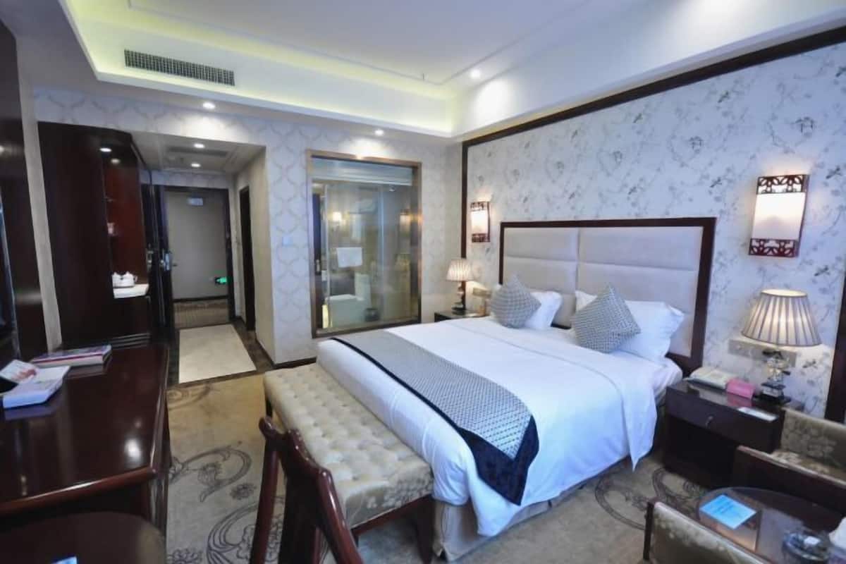 A hotel room with a large bed, a desk, a chair, a bench, a bedside table, and a lamp.