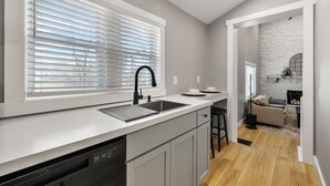 A modern kitchen with a built-in dishwasher, a large window with blinds, and a stone accent wall. - Hoosier HQ: Sleek 3-Bed Near IU (Bloomington)