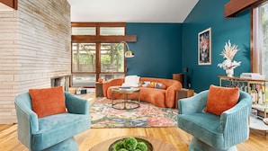 A modern living room with a brick wall, a large window, and a teal sofa with orange cushions.