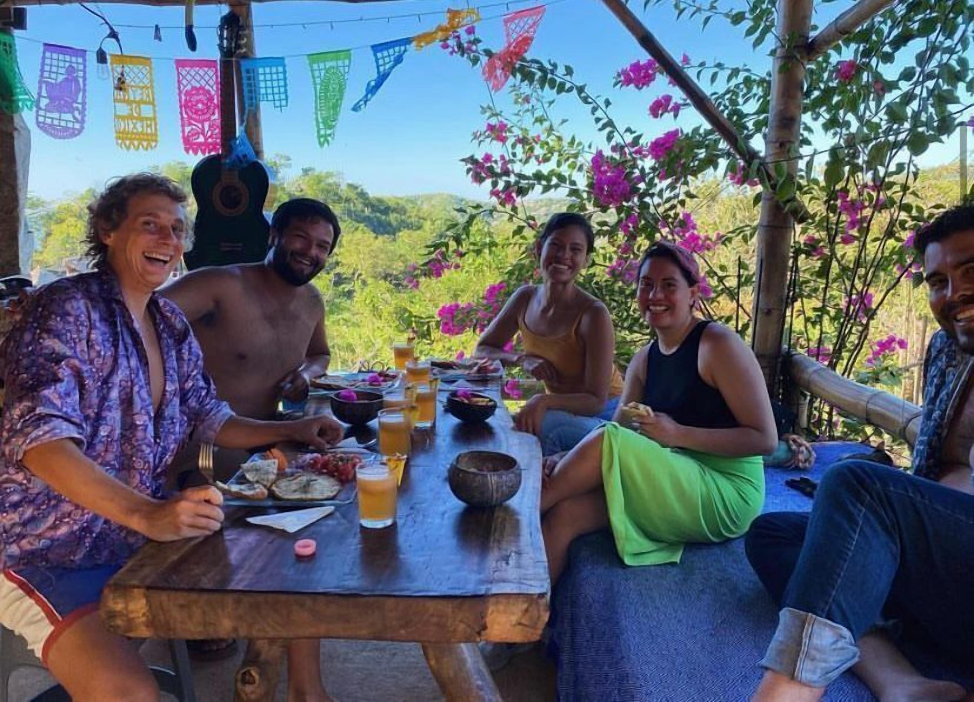 A group of people dining outdoors at a wooden table with a view of a mountainous landscape.