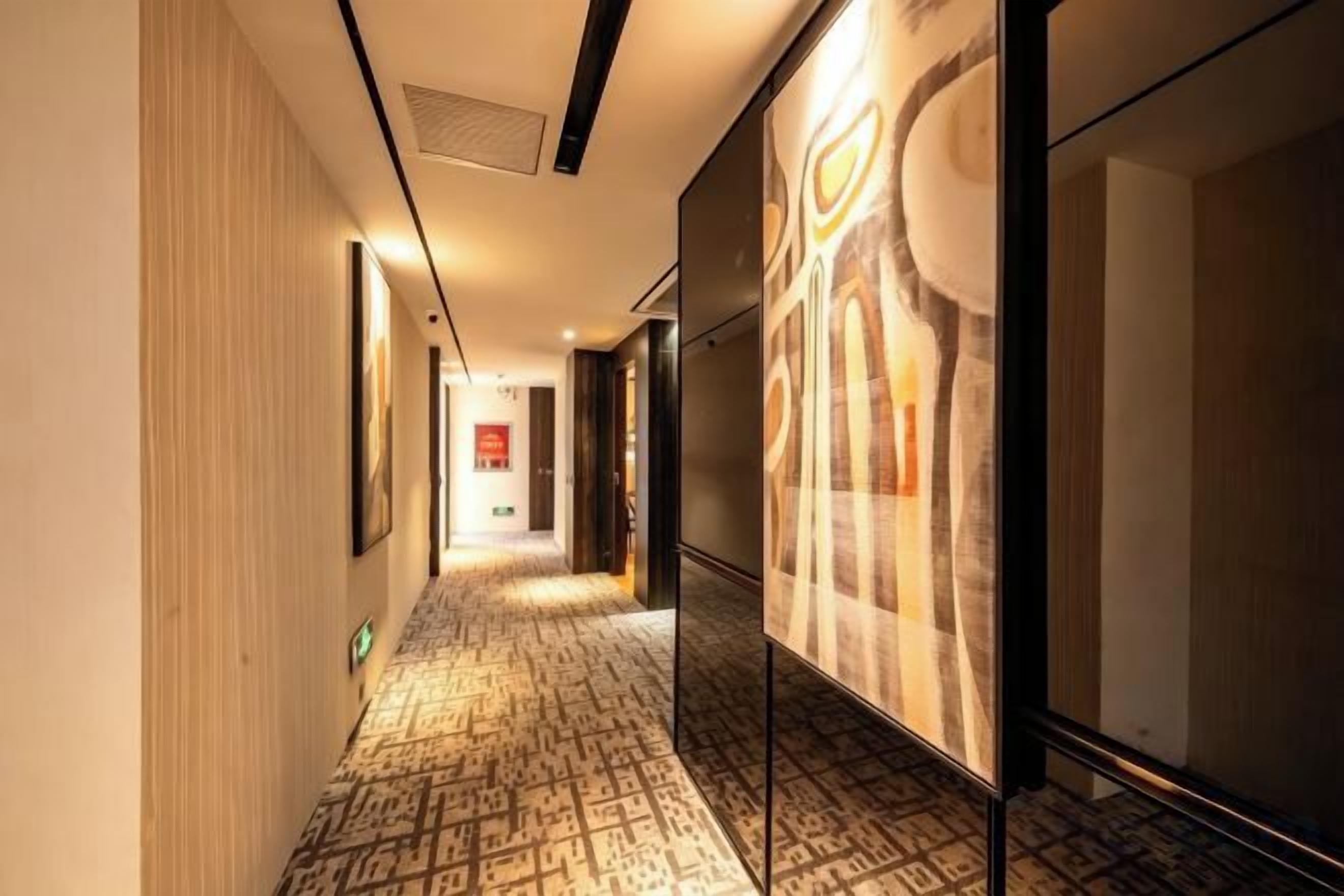 A hotel corridor with patterned carpet, framed artwork on the walls, and a door at the end.