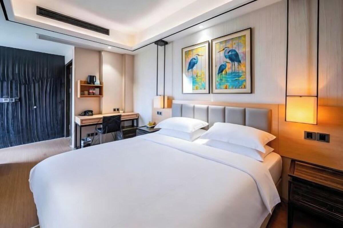 Une chambre d’hôtel moderne avec un grand lit, un bureau et une chaise. Deux tableaux sont accrochés au mur, au-dessus du lit.