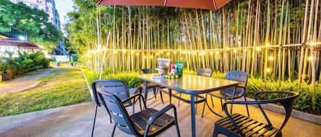 An outdoor seating area with a table and chairs under a large umbrella, surrounded by bamboo plants and string lights.