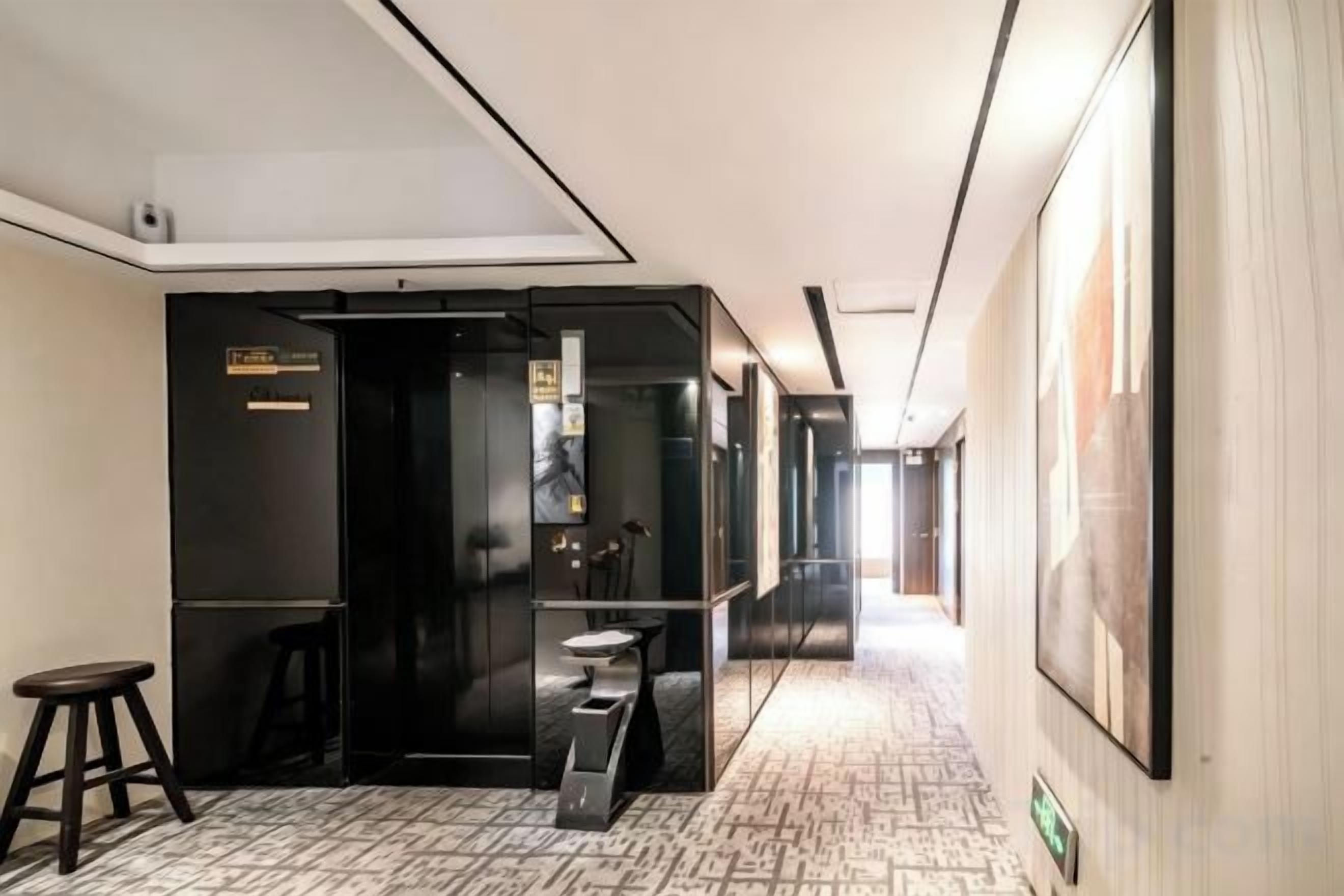 Een hotelgang met een zwarte lift, een tafeltje met een stoel, en een groot fotokader aan de muur.