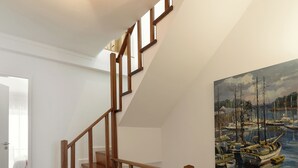 Un escalier en bois, doté d’une rampe, mène à une pièce où est accrochée une peinture représentant un paysage portuaire.