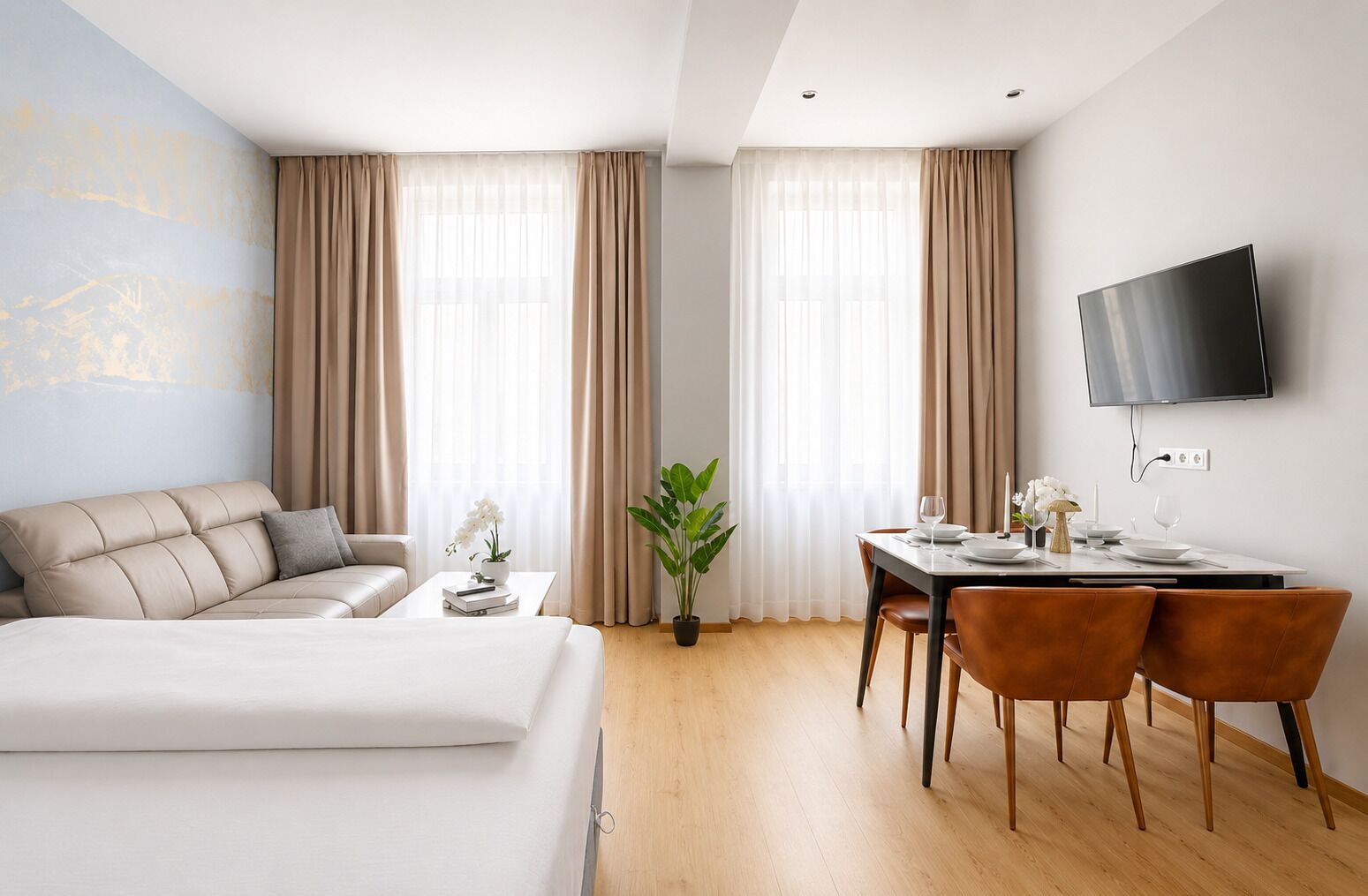 A hotel room with a bed, a dining table with chairs, a TV mounted on the wall, and large windows with curtains.