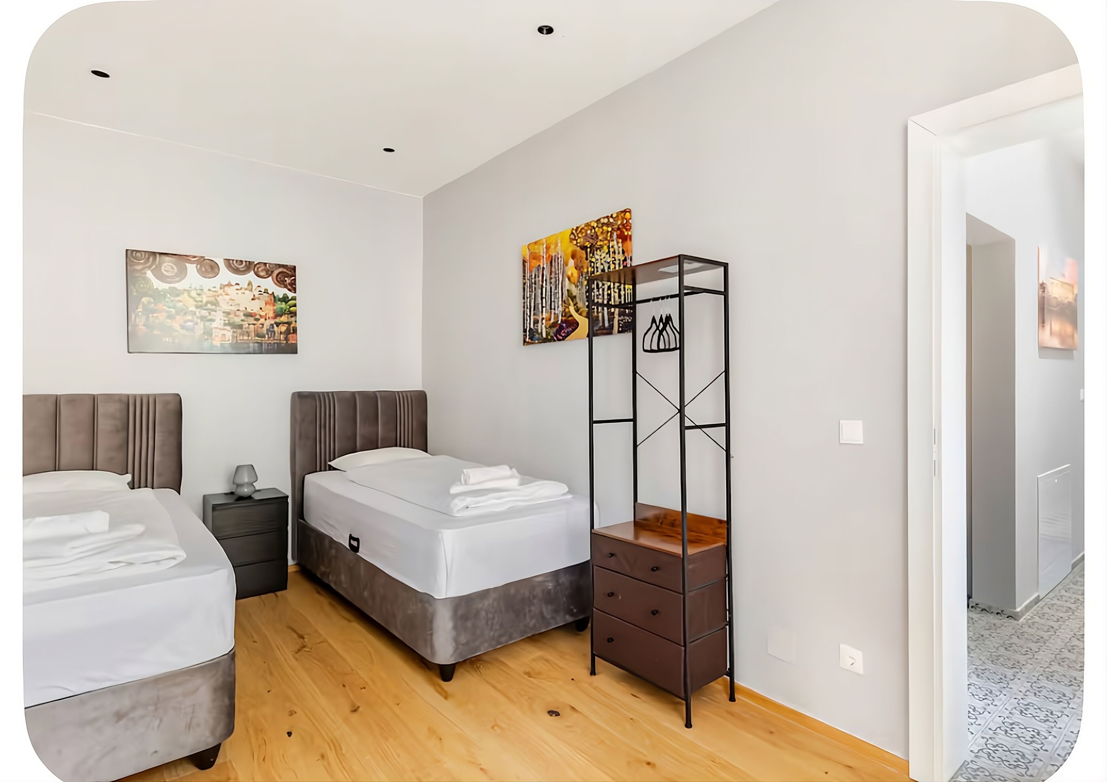 A hotel room with two beds, a wooden floor, a metal storage unit, and artwork on the walls.