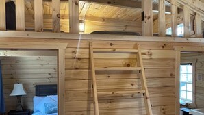 Interior de madera con un dormitorio en un entrepiso, una escalera que lleva al área de descanso superior, una cama con almohada y una mesita de noche con lámpara.