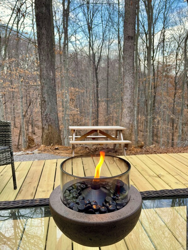 A glass-top fire pit with a visible flame, surrounded by a deck with a wooden picnic table in the background.