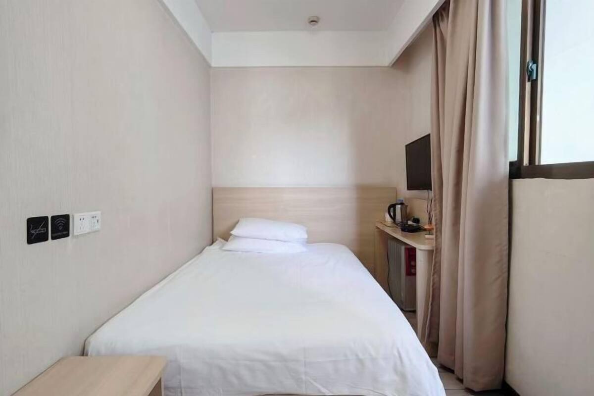 Una habitación de hotel pequeña y limpia con una cama individual, una mesita de noche, un televisor y una ventana con cortinas.