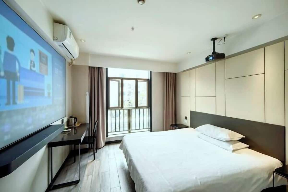 Una habitación de hotel con cama, escritorio, pantalla para proyector y ventana con cortinas.