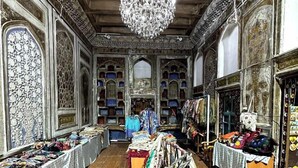 An interior with ornate wooden paneling, a chandelier, and various textiles displayed on tables. - Shapira Boutique Hotel (Buxoro)