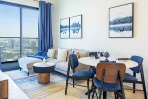 A modern living room with a sofa, coffee table, and dining area with chairs. There are two framed pictures on the wall, a large window with curtains, and a view of a cityscape.