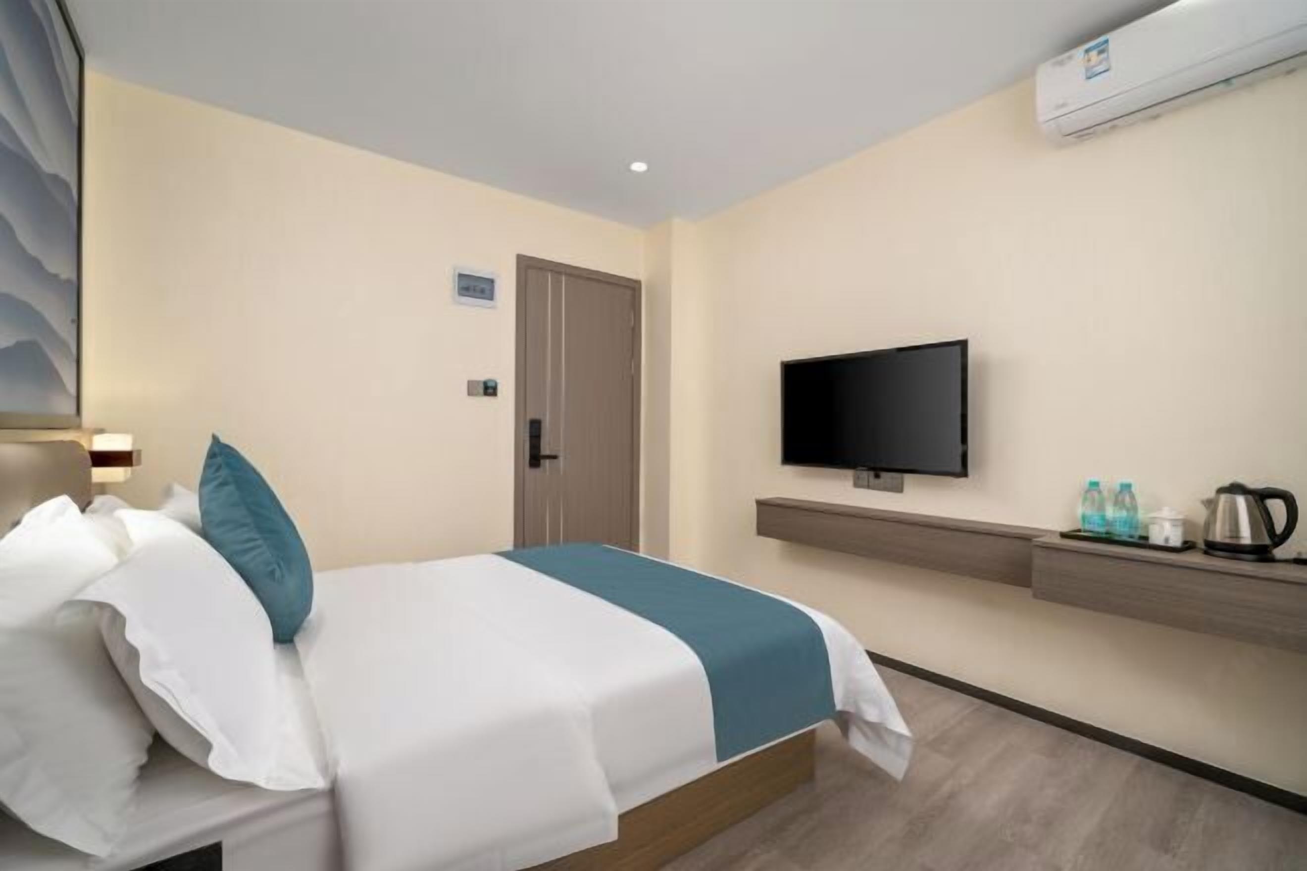 Ein Hotelzimmer mit Bett, Fernseher, Tür und einem an der Wand befestigten Regal mit Wasserkocher und Wasserflaschen.