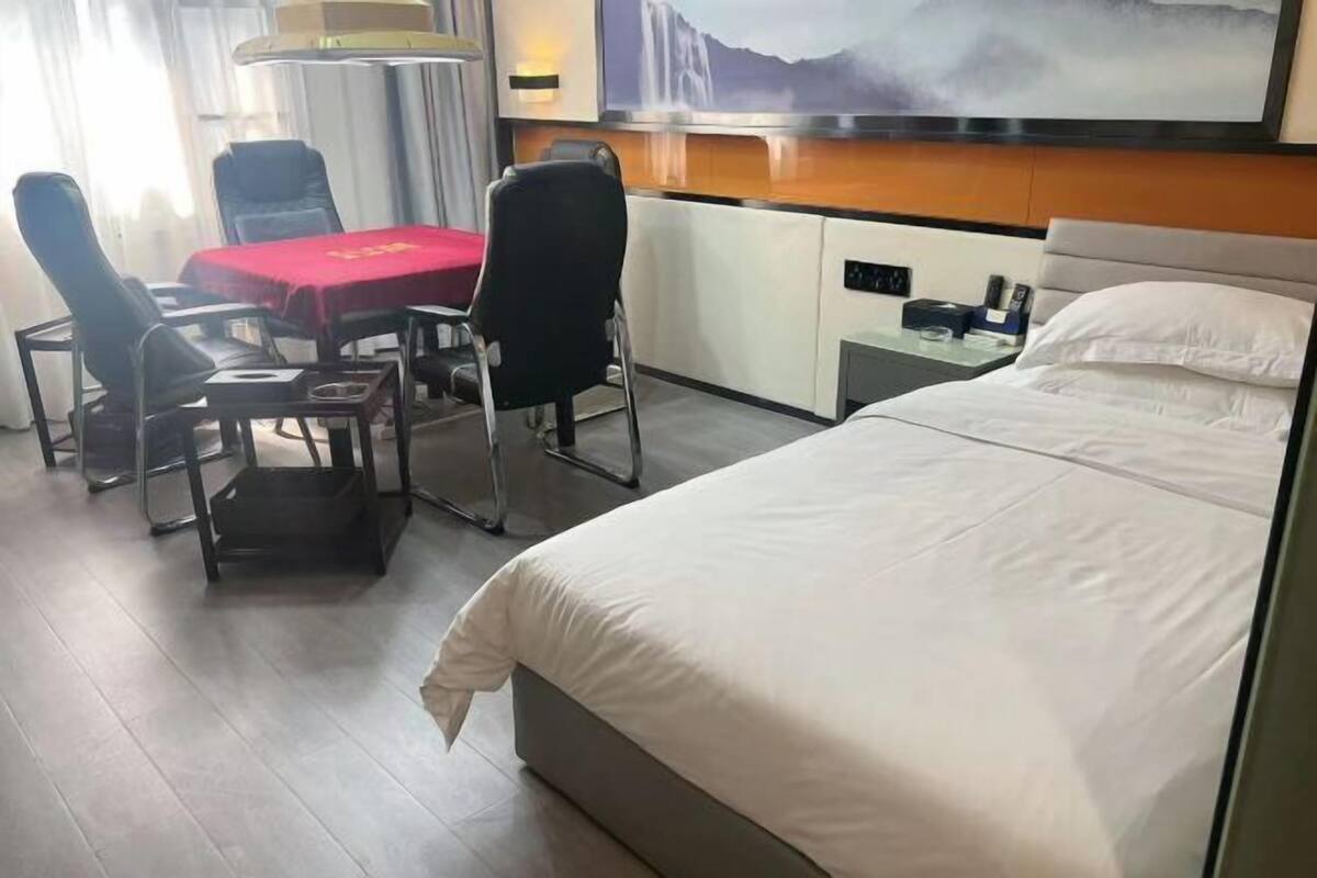 Una habitación de hotel con una cama, un escritorio con mantel rojo, dos sillas, una lámpara y un cuadro en la pared.