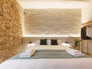 A bedroom with a brick wall, a bed with white linens, a green throw, and a wooden wardrobe with clothes. - Bravissimo Estudi Verd; Spacious 3-bedroom apartment in the Historic Centre (Girona)