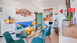 A modern living room with a glass dining table, teal chairs, a blue sofa, and a kitchen in the background. - [Spacious +sea view with garden] (Arenzano)