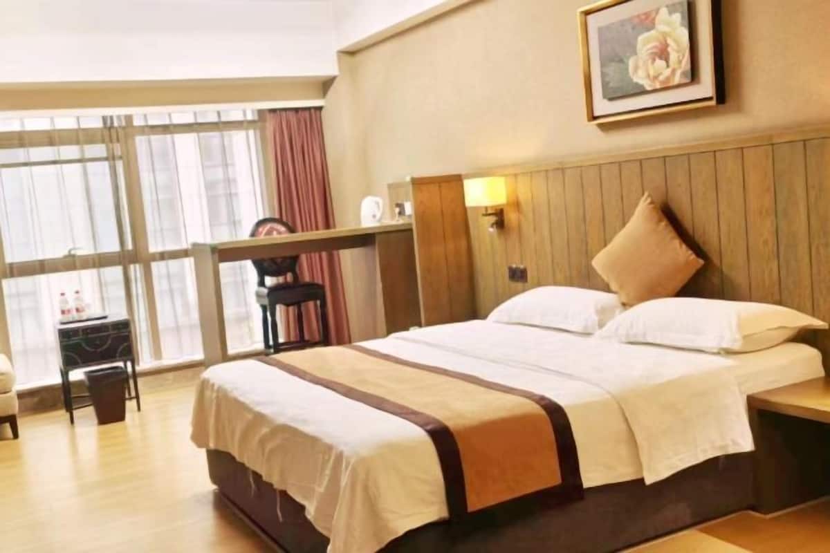 Une chambre d’hôtel avec un grand lit, une tête de lit en bois, une table de chevet, une commode, un bureau avec une chaise, une fenêtre avec des rideaux et un tableau encadré au mur.
