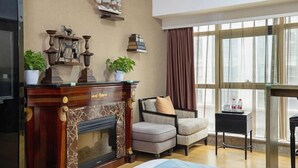 Ein Zimmer mit Kamin, zwei Stühlen, einem kleinen Tisch und einem großen Fenster mit Vorhängen.