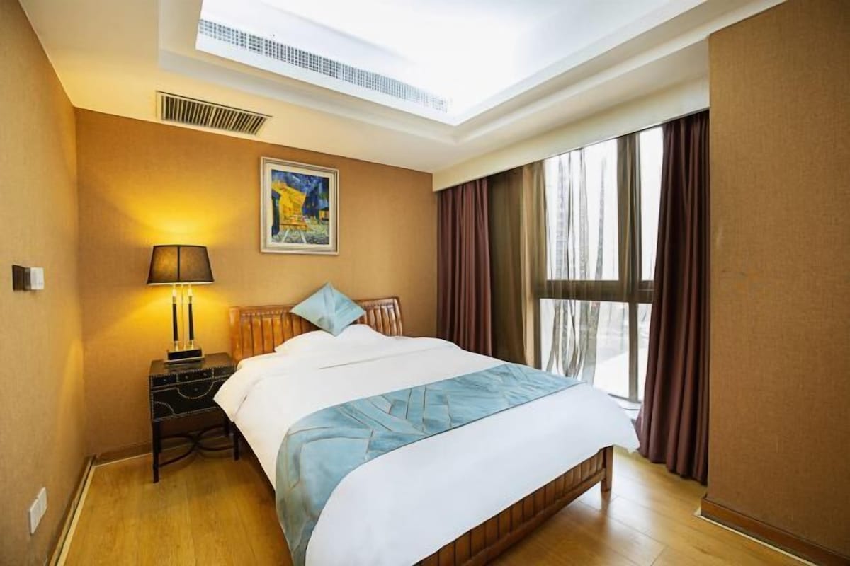 A hotel room with a large bed, a nightstand with a lamp, a painting on the wall, and a window with curtains.