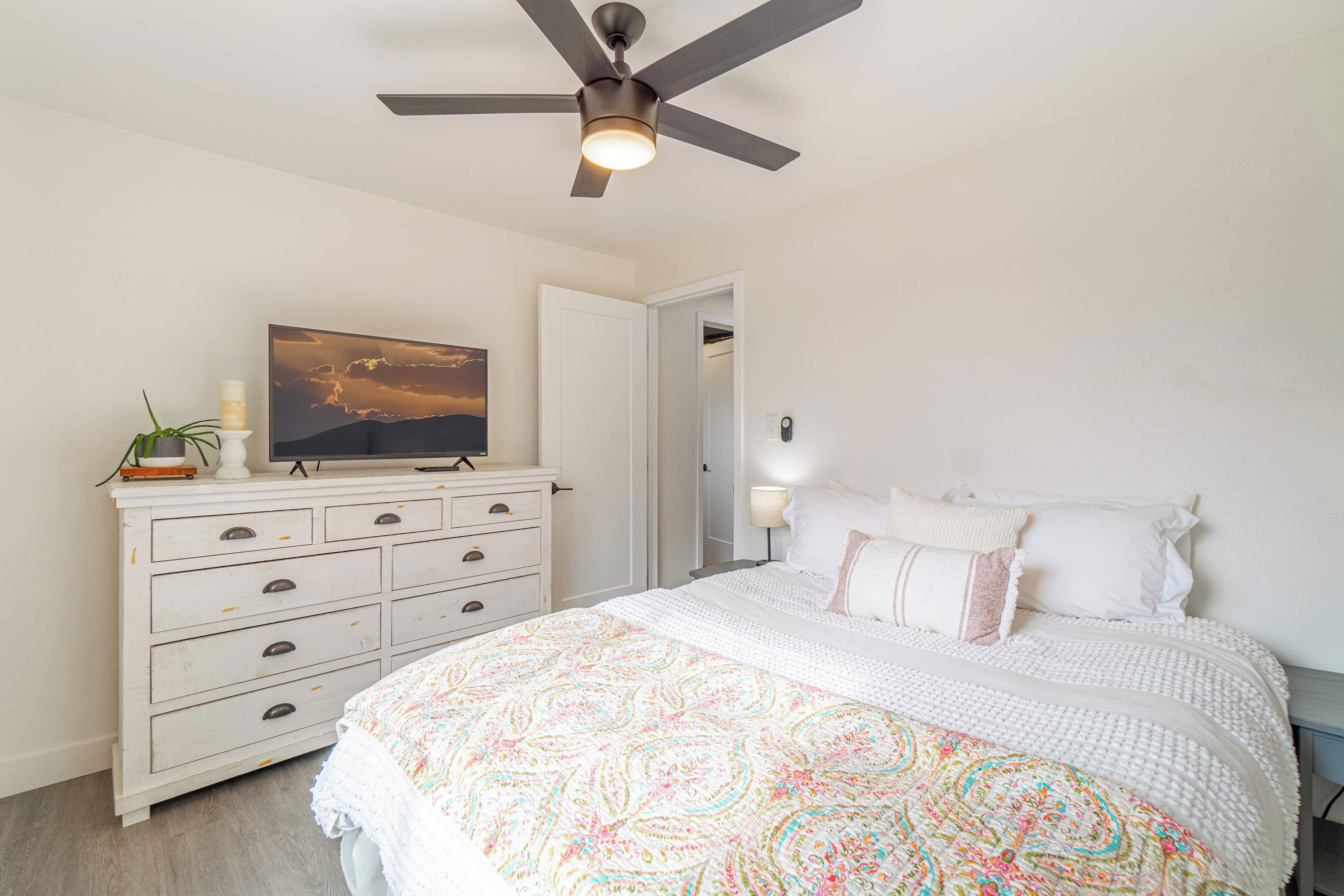 Un dormitorio con cama, cómoda, ventilador de techo, un cuadro y mesita de noche con lámpara.