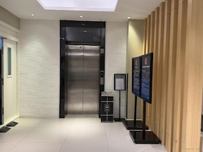 A modern elevator with a metallic finish, a reception area with a wooden panel wall, and a digital information board.