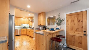 A kitchen with wooden cabinets, a stainless steel refrigerator, a bar area with stools, and a large wooden door.