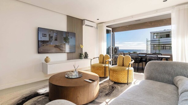 Een moderne woonkamer met een ronde salontafel, een bank en gele fauteuils met een patroon.