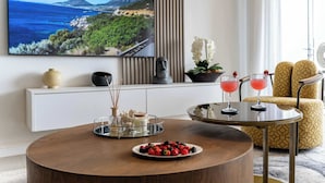 Een moderne woonkamer met een ronde houten salontafel, een televisie waarop een idyllisch landschap te zien is, een geel gestreept bankstel en twee drankjes op een bijzettafeltje.