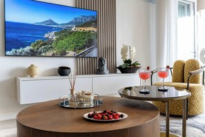 A modern living room with a round wooden coffee table, a television displaying a scenic landscape, a yellow patterned sofa, and two drinks on a small table.