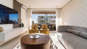 Een moderne woonkamer met een flatscreen televisie, een ronde houten salontafel, gele gestreepte stoelen, een grijze bank en een uitzicht op een stadsgezicht door grote ramen.