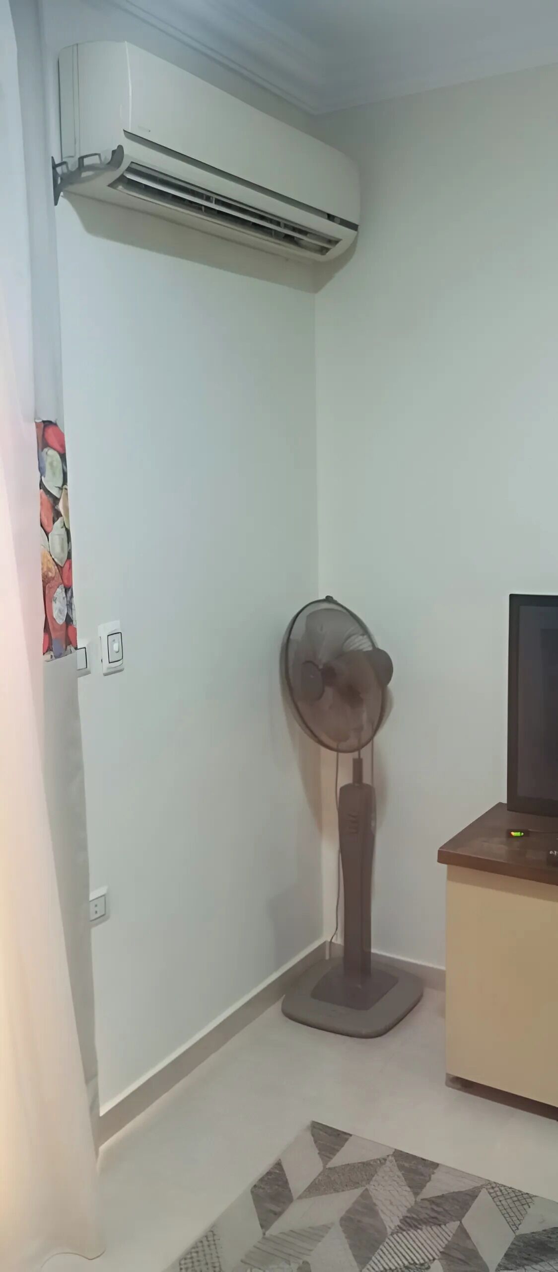 Una habitación de esquina con aire acondicionado, un ventilador de pie, un televisor sobre un mueble de madera y una alfombra estampada.