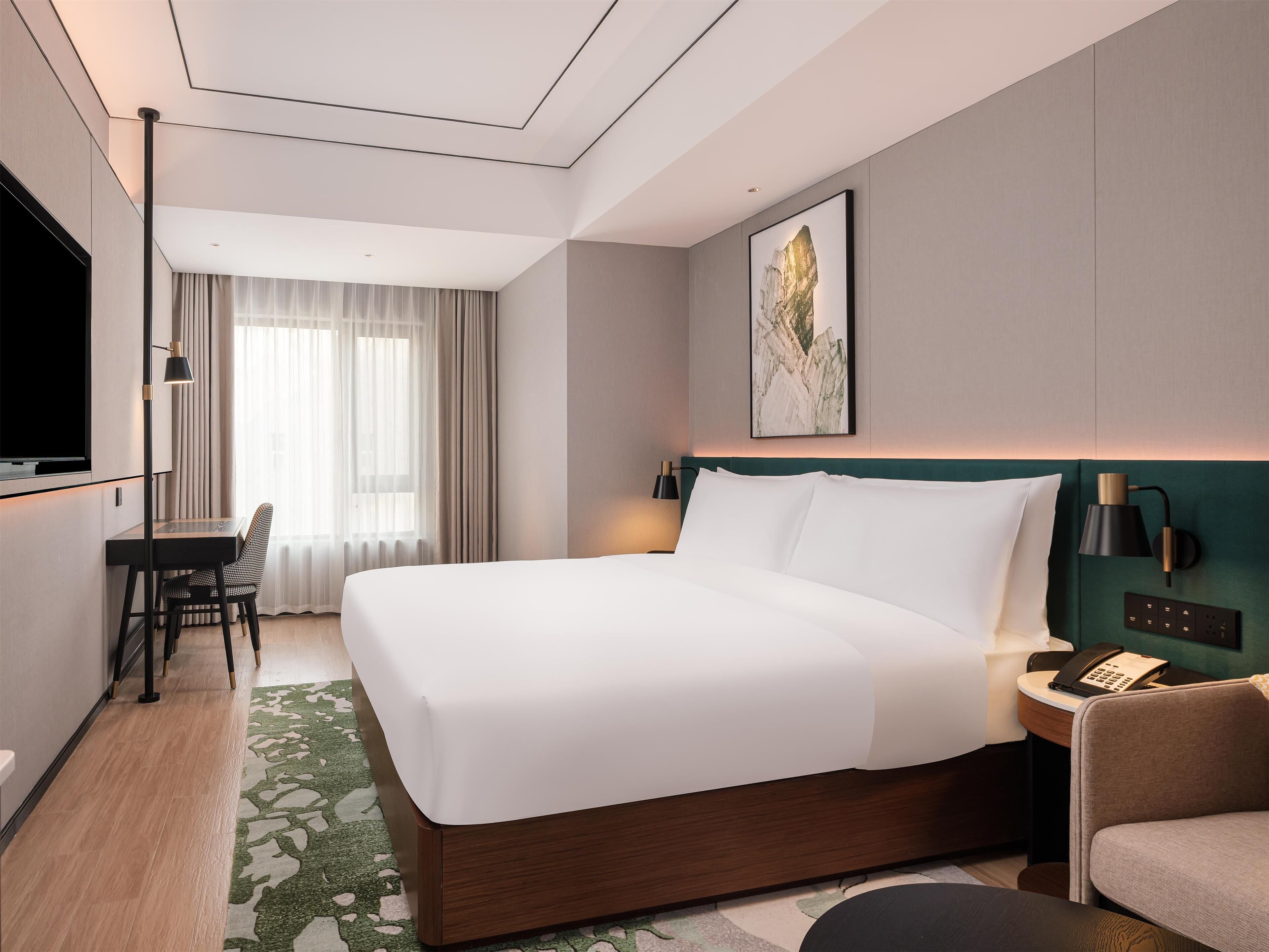 A modern hotel room with a large bed, a desk, a chair, a TV, and a painting on the wall.