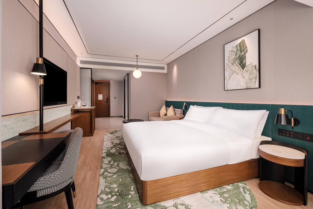 A modern hotel room with a large bed, a desk with a chair, a TV, and a bedside table with a lamp.