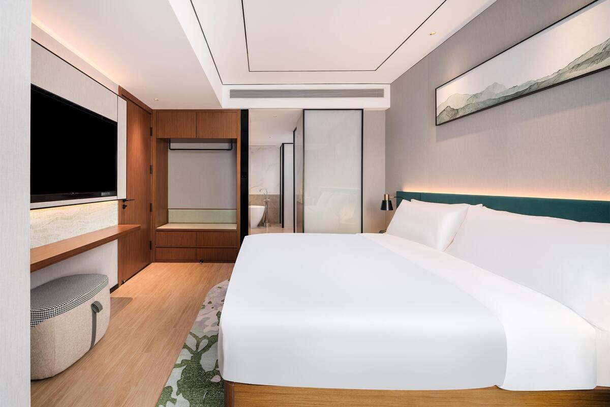 A modern hotel room with a large bed, a flat-screen TV, a wooden cabinet, and a bathroom visible through a glass door.